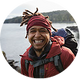 A close-up portrait of a smiling man wearing a red patterned headwrap and a red jacket, with a backpack visible on his shoulders, set against an outdoor coastal background.
