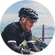 A man wearing a black cycling helmet and a black cycling jersey with white stripes, riding a bicycle against a blurred outdoor background with a bridge and water visible.