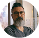 A circular portrait of a man with glasses, a full beard, and a dark gray t-shirt, set against a light gray background.